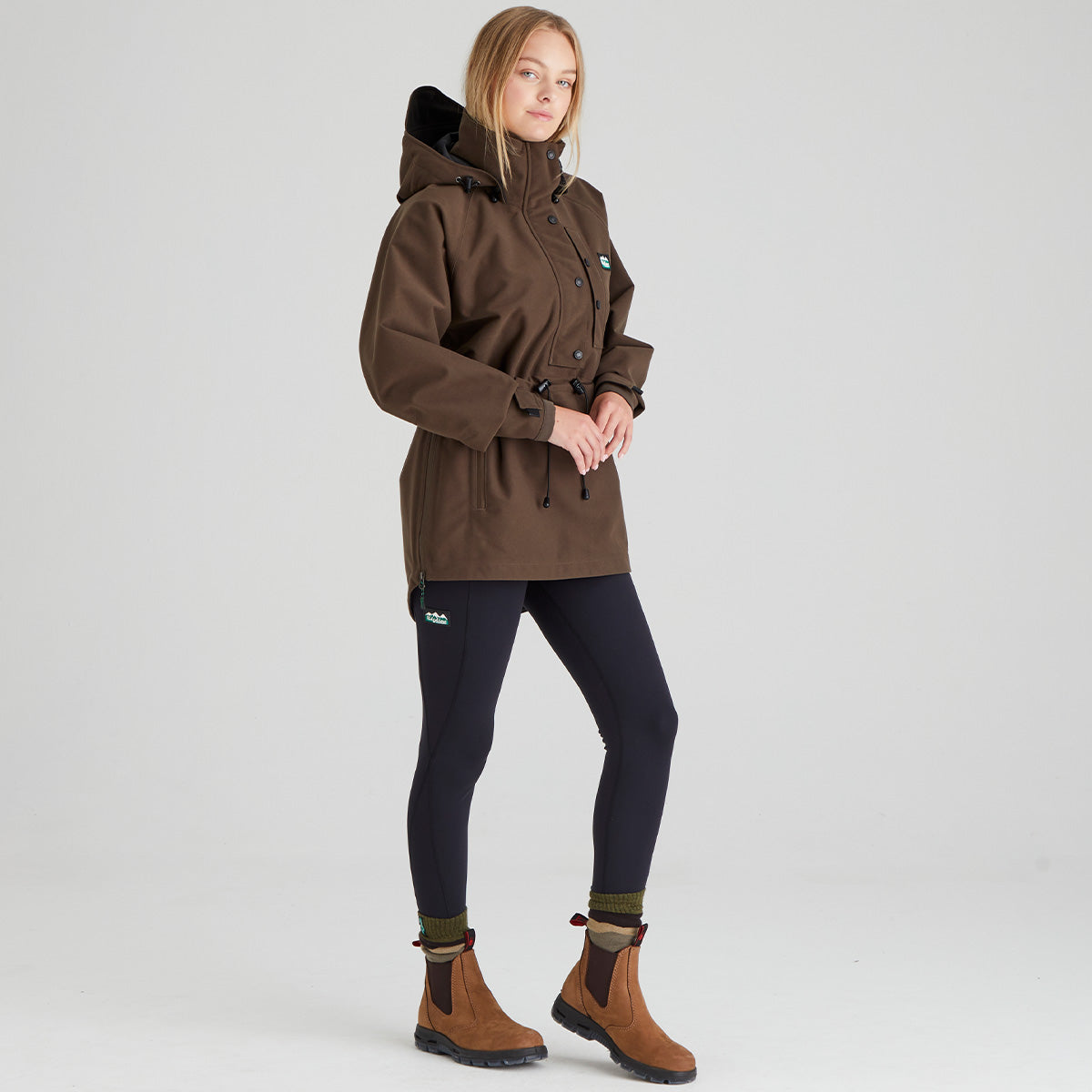 Woman wearing a brown waterproof jacket, black leggings, and brown boots on a light grey background
