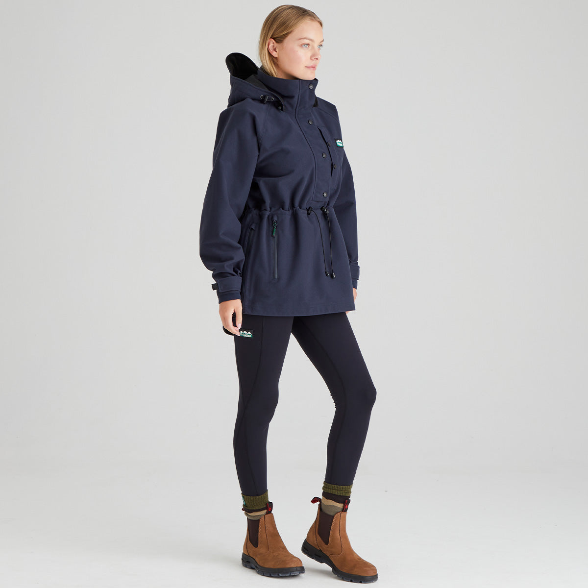Woman wearing a navy blue waterproof jacket, leggings, and brown boots on a light grey background