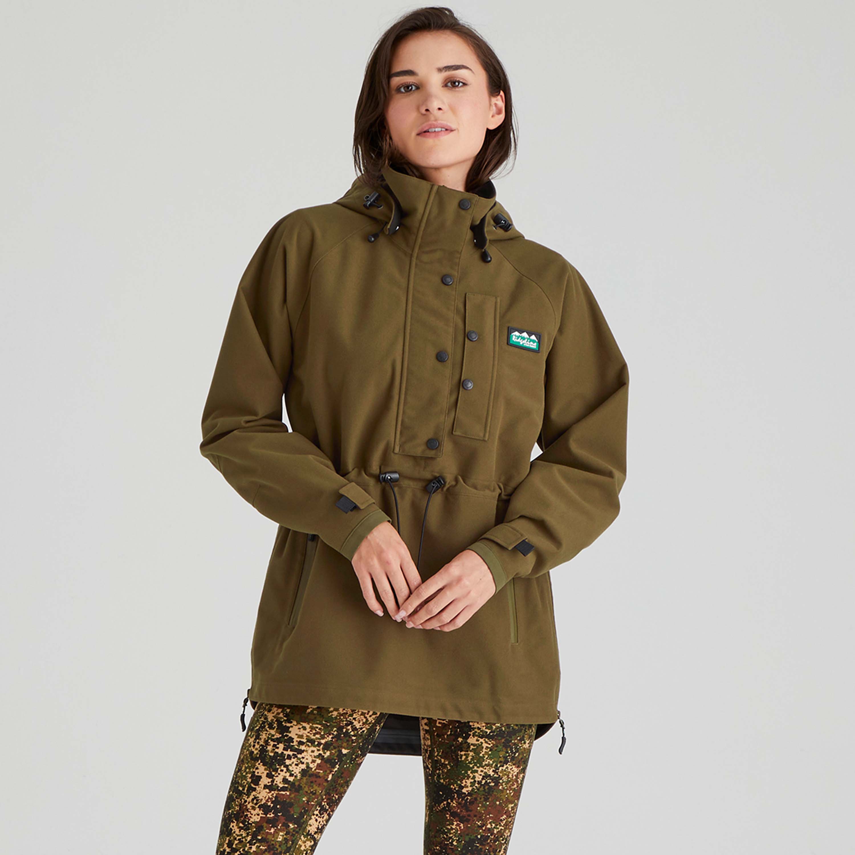 Person wearing a green/brown jacket with a brand logo on a light grey background