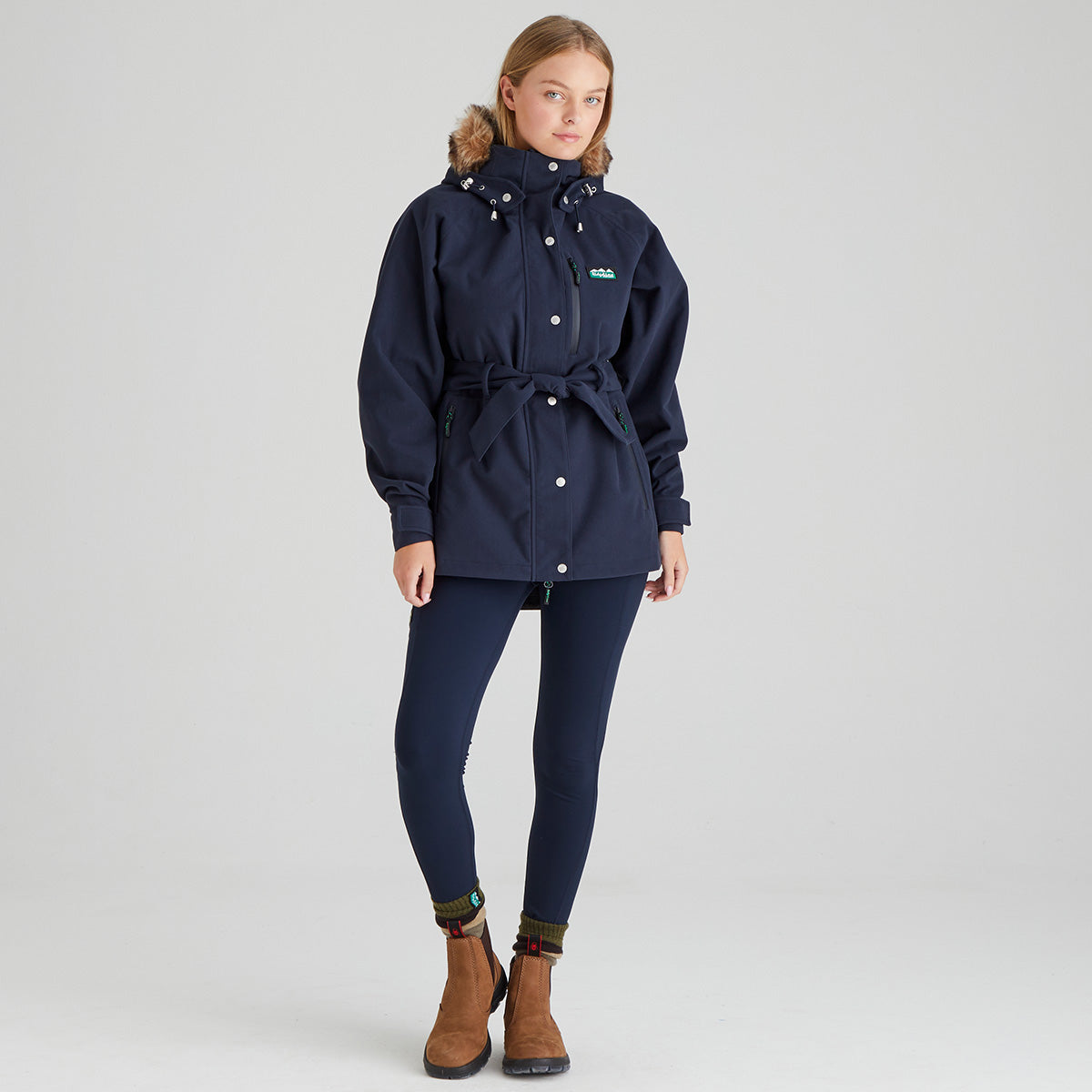 Person wearing a navy blue waterproof jacket and pants with brown boots on a light grey background