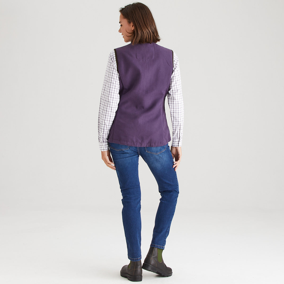 Person wearing a purple vest over a checkered shirt with blue jeans on a white background