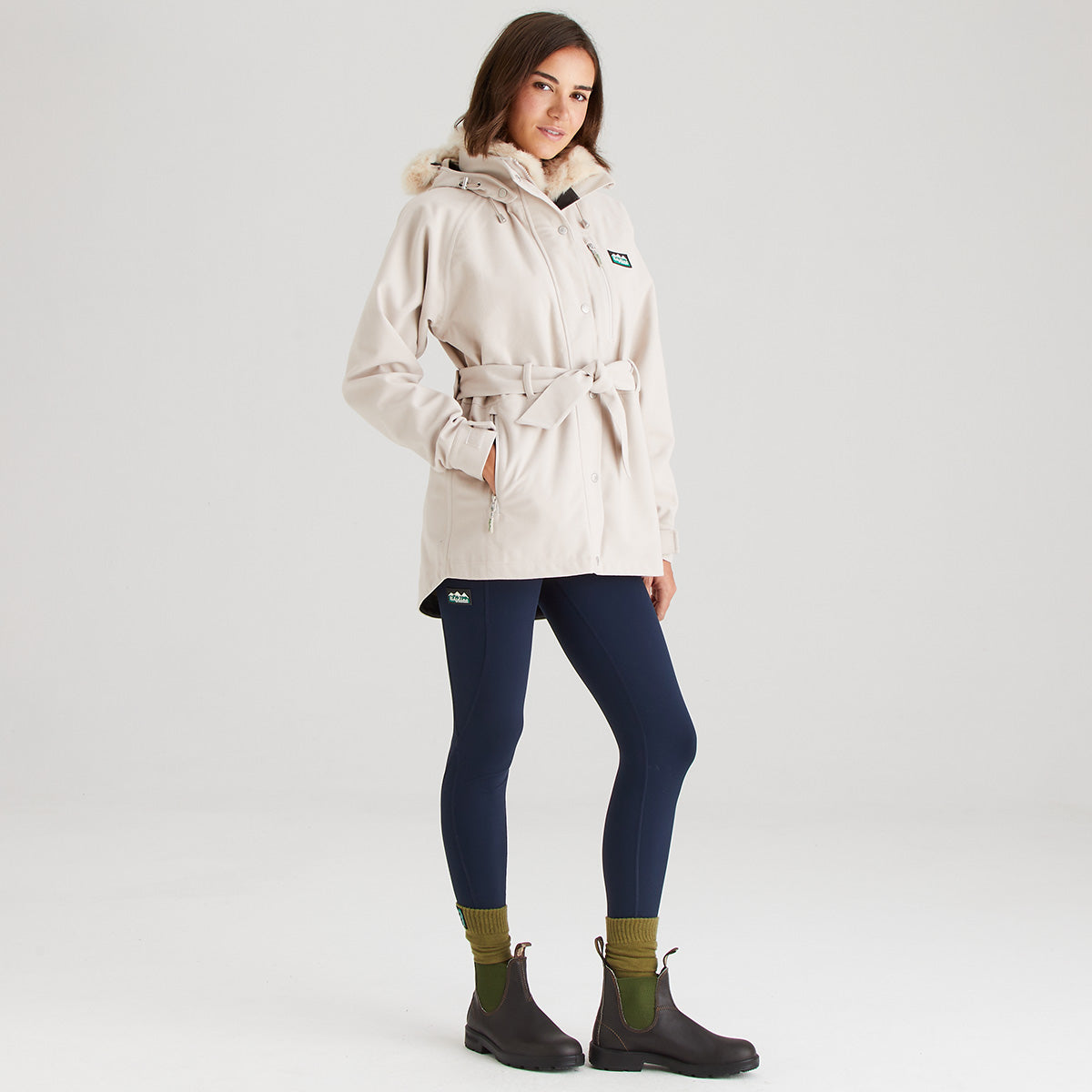 Woman wearing a cream coat, navy leggings, and black boots on a white background