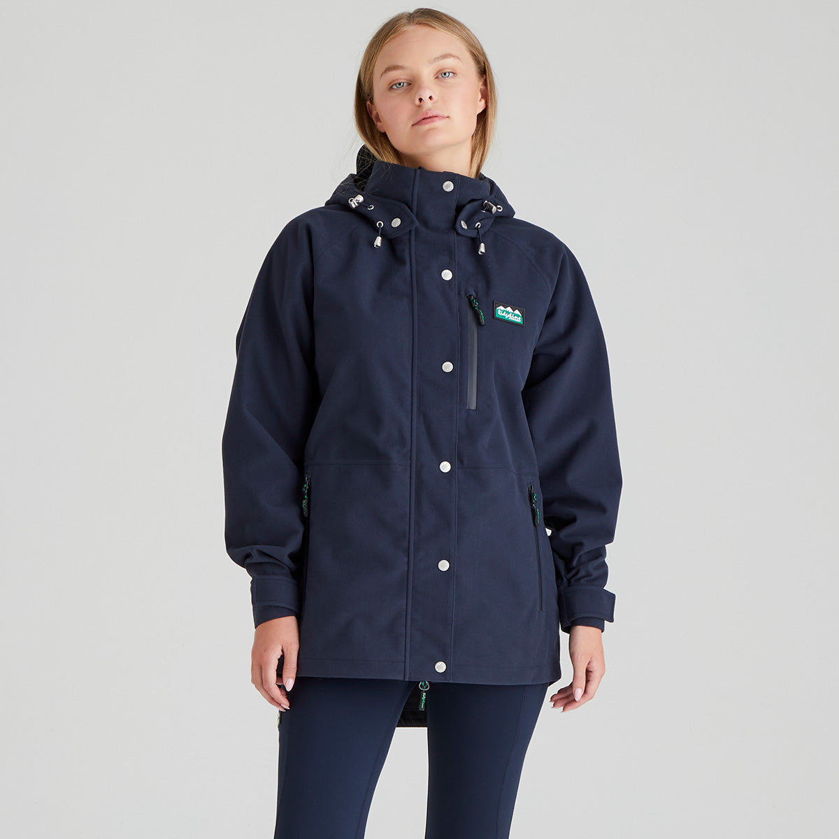 A woman wearing a navy blue hooded rain jacket with white snap buttons and a small green and white logo on the left chest, paired with matching navy pants, against a light gray studio background.