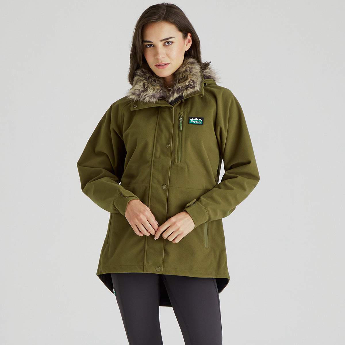 A woman wearing an olive green hooded parka jacket with a faux-fur trimmed hood and a small brand patch on the chest, photographed against a light gray studio background.