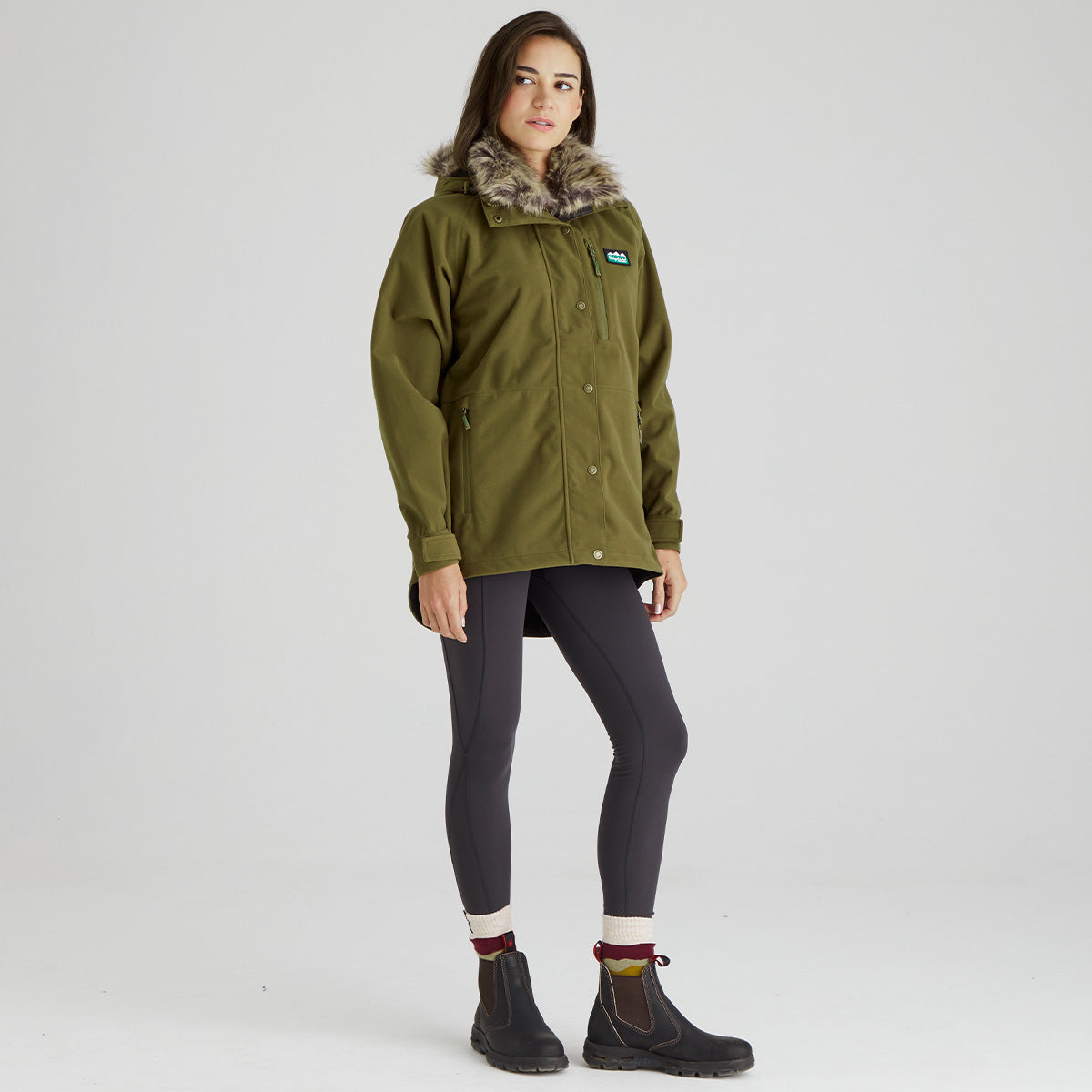A woman modeling an olive green outdoor jacket with a brown faux-fur trimmed hood, paired with dark gray leggings and black boots, against a light gray studio background.