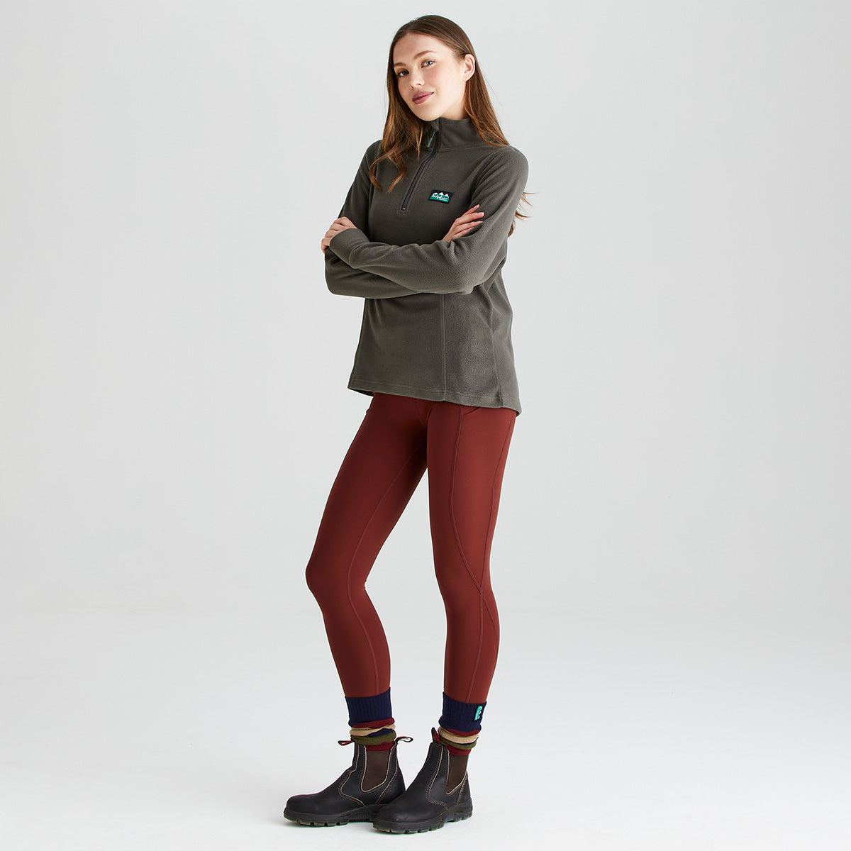 Full-length studio image of a person wearing a grey Ridgeline quarter-zip fleece pullover, rust-red performance leggings, striped socks, and black ankle boots. The fleece features a high stand-up collar, a quarter-zip neckline, long sleeves, and a Ridgeline logo patch on the left chest. The leggings have a high-waisted fit with contoured seam detailing for a streamlined silhouette. Photographed against a plain light-grey background, the image highlights the comfortable outdoor layering combi