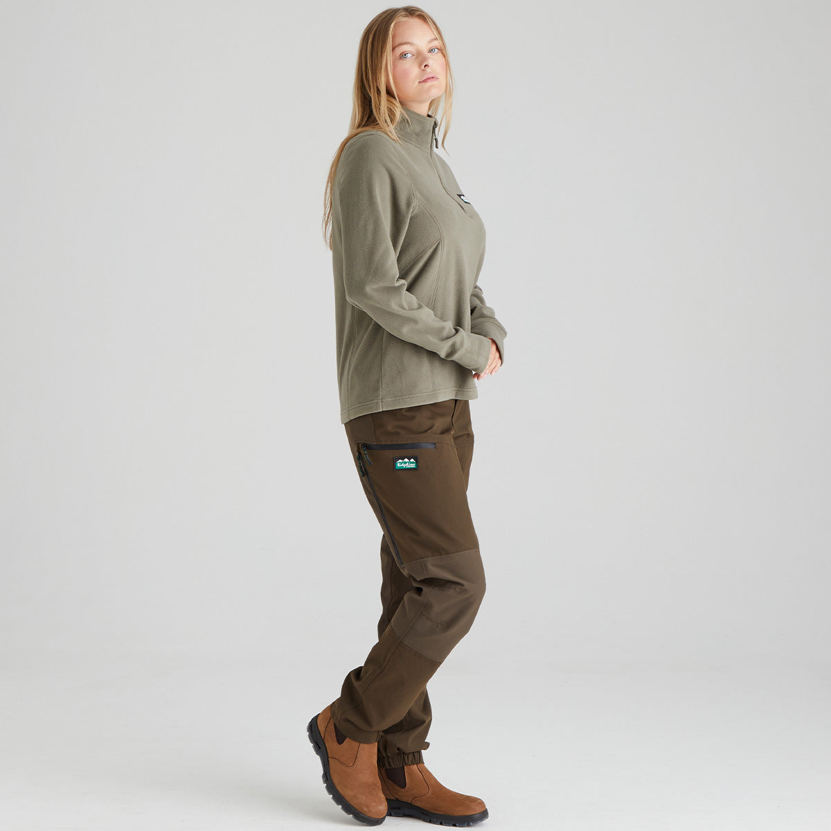 Person wearing a green long-sleeve fleece top and brown pants on a light grey background