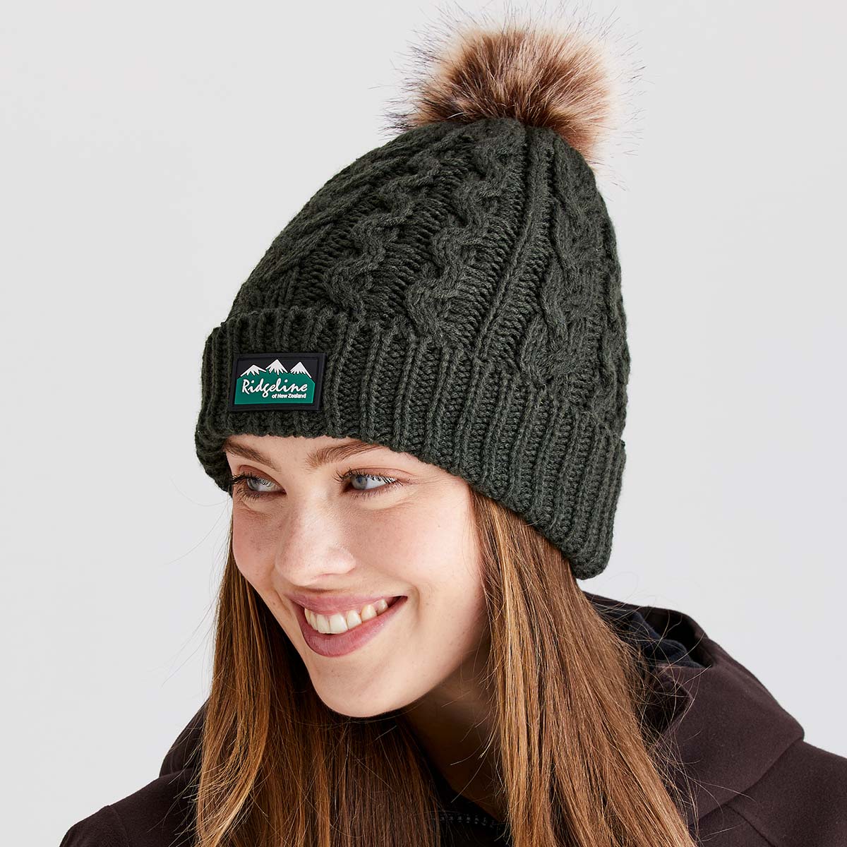 Lifestyle image showing a person wearing a dark charcoal cable-knit beanie with a ribbed cuff and a large faux-fur pom-pom on the crown. A Ridgeline of New Zealand logo patch is attached to the front cuff. The beanie features a textured cable-knit pattern throughout, providing a classic winter look. The person is wearing a dark hooded jacket and is photographed against a light grey background, highlighting the beanie's warm knit construction, decorative pom-pom, and outdoor-inspired style.
