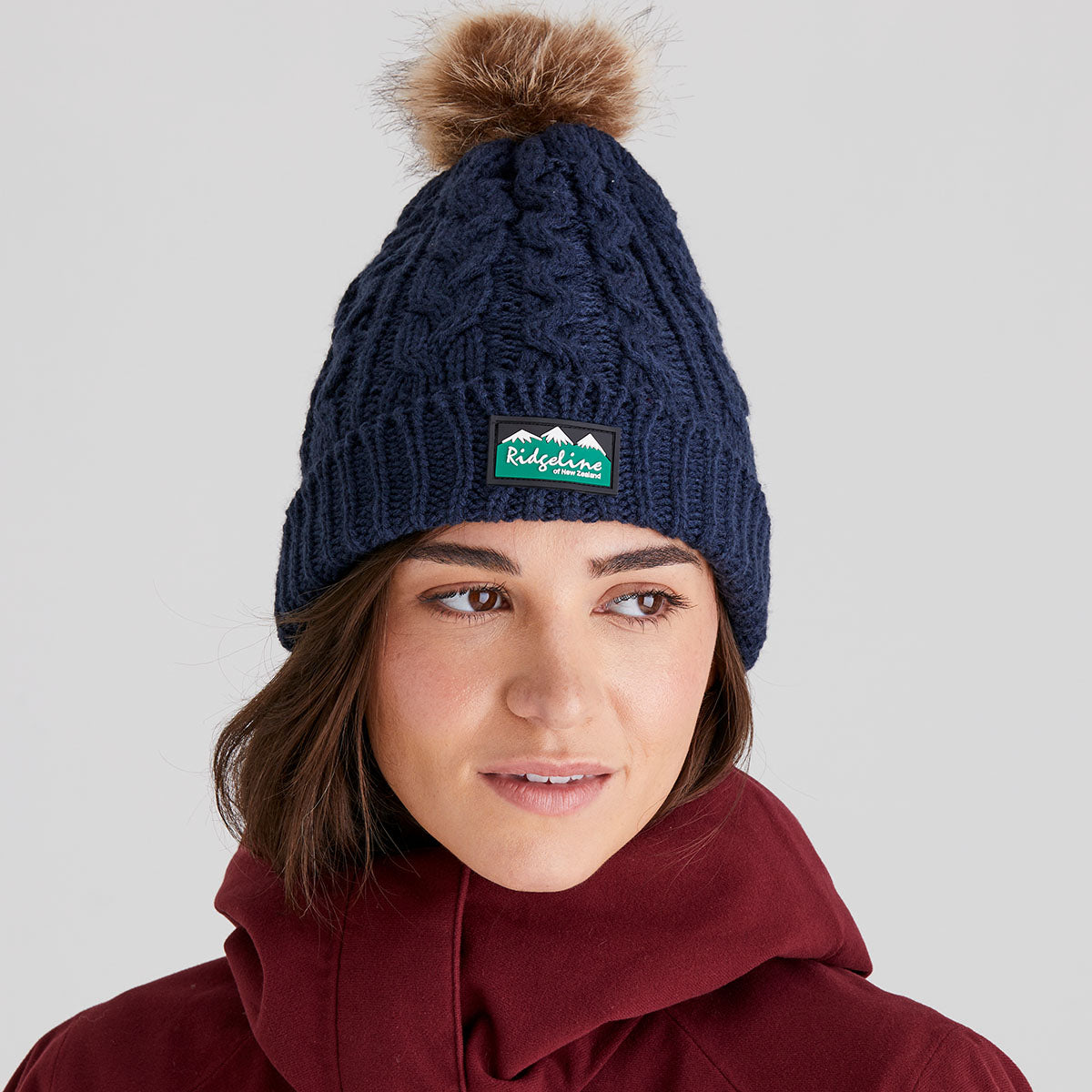 Woman wearing a navy knit beanie with a pom-pom and a visible ridgeline clothing brand logo on a light grey background.
