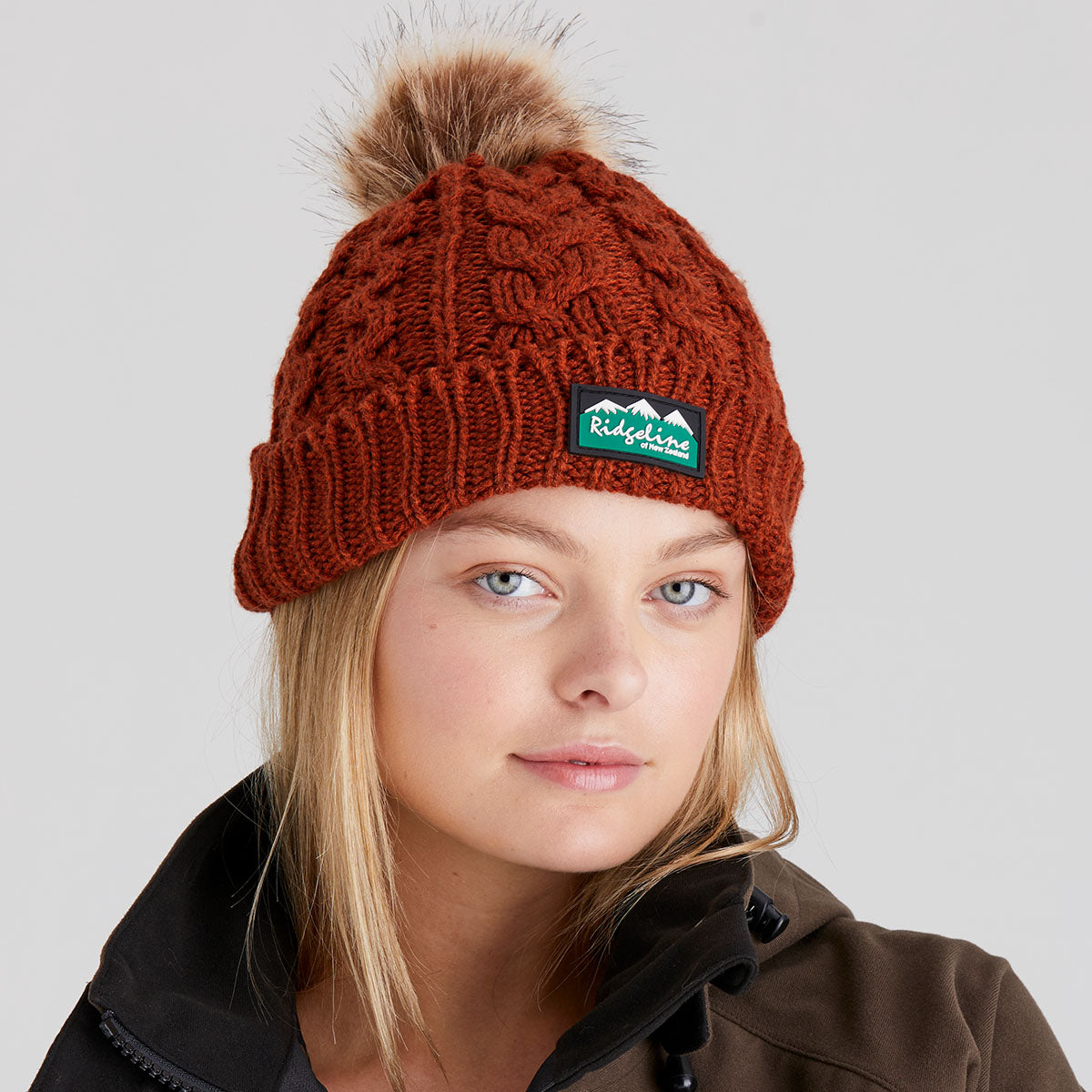 woman wearing a orange knit beanie with a pom-pom and a visible brand logo on a light grey background