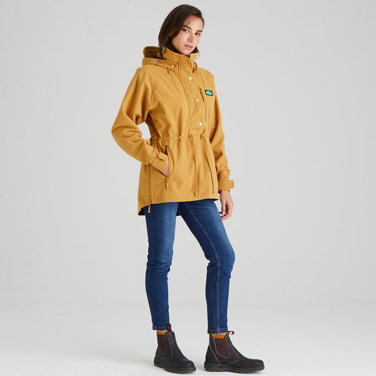 Woman wearing a mustard yellow jacket with a brand logo on a light gray background