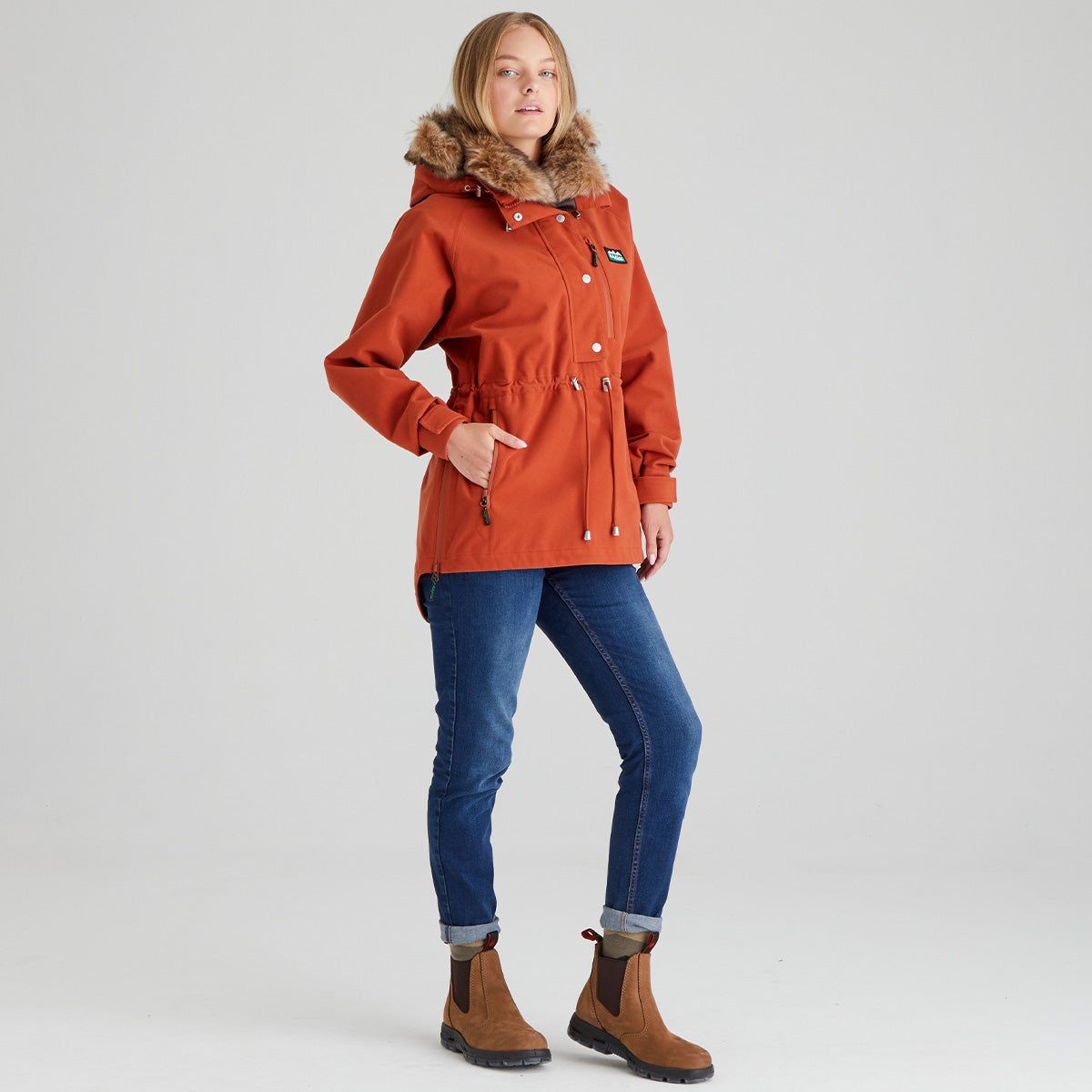 Person wearing an orange coat with fur hood, blue jeans, and brown boots on a light grey background