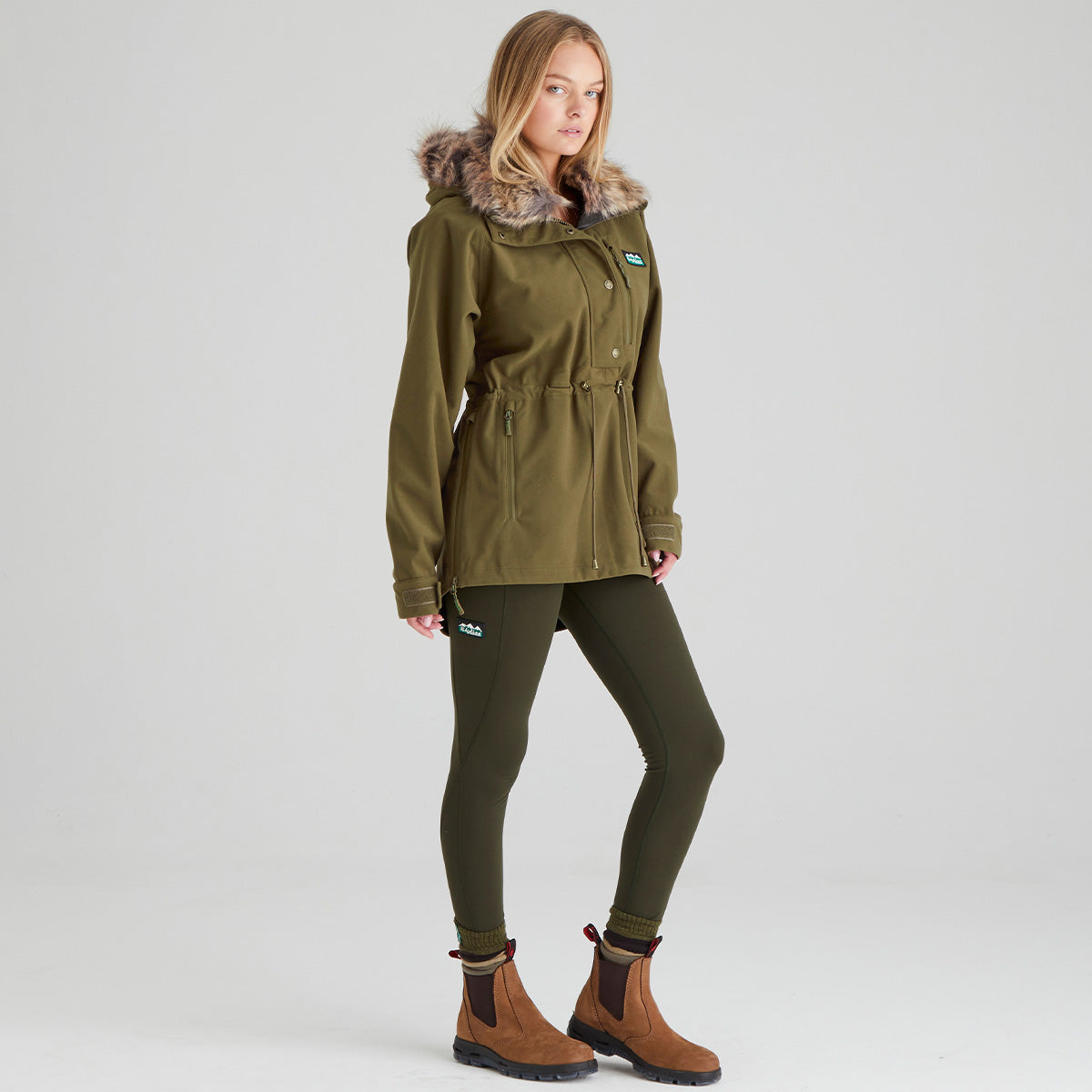Woman wearing a green parka, leggings, and boots on a light grey background