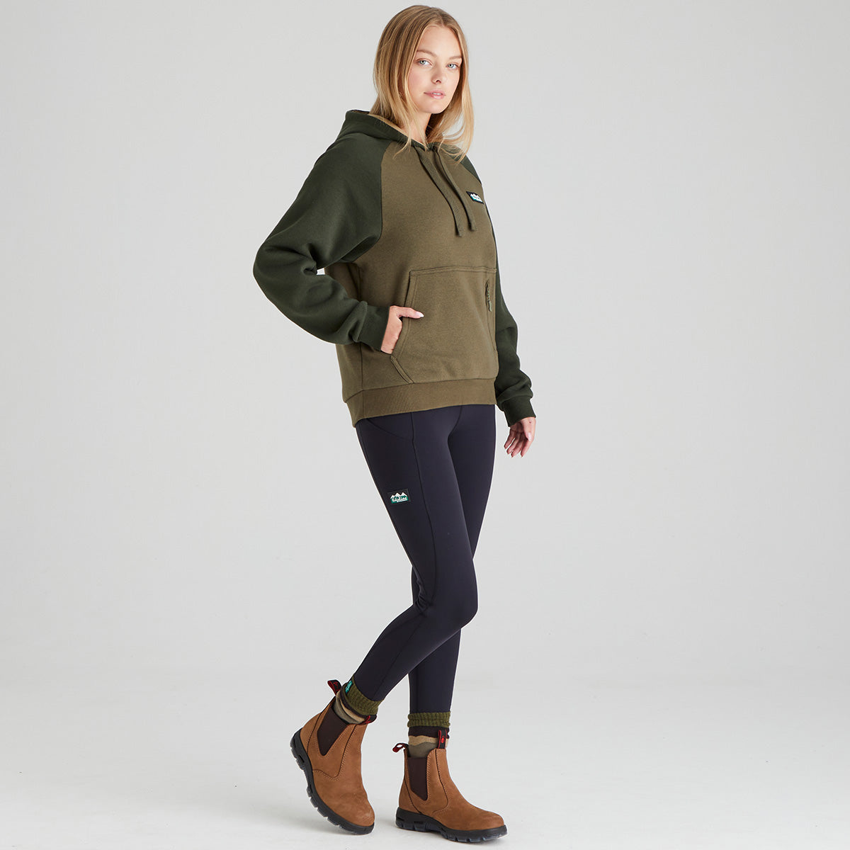 Woman wearing a green hoodie and dark leggings with a logo on a light grey background