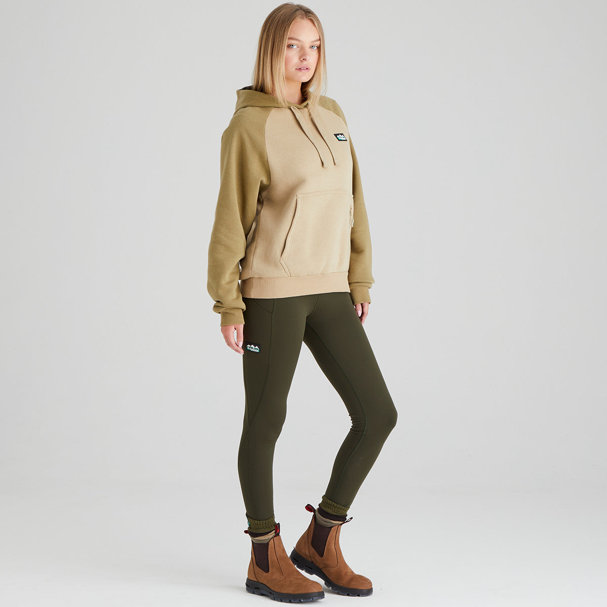 Person wearing a beige hoodie and green leggings with brown boots on a light grey background