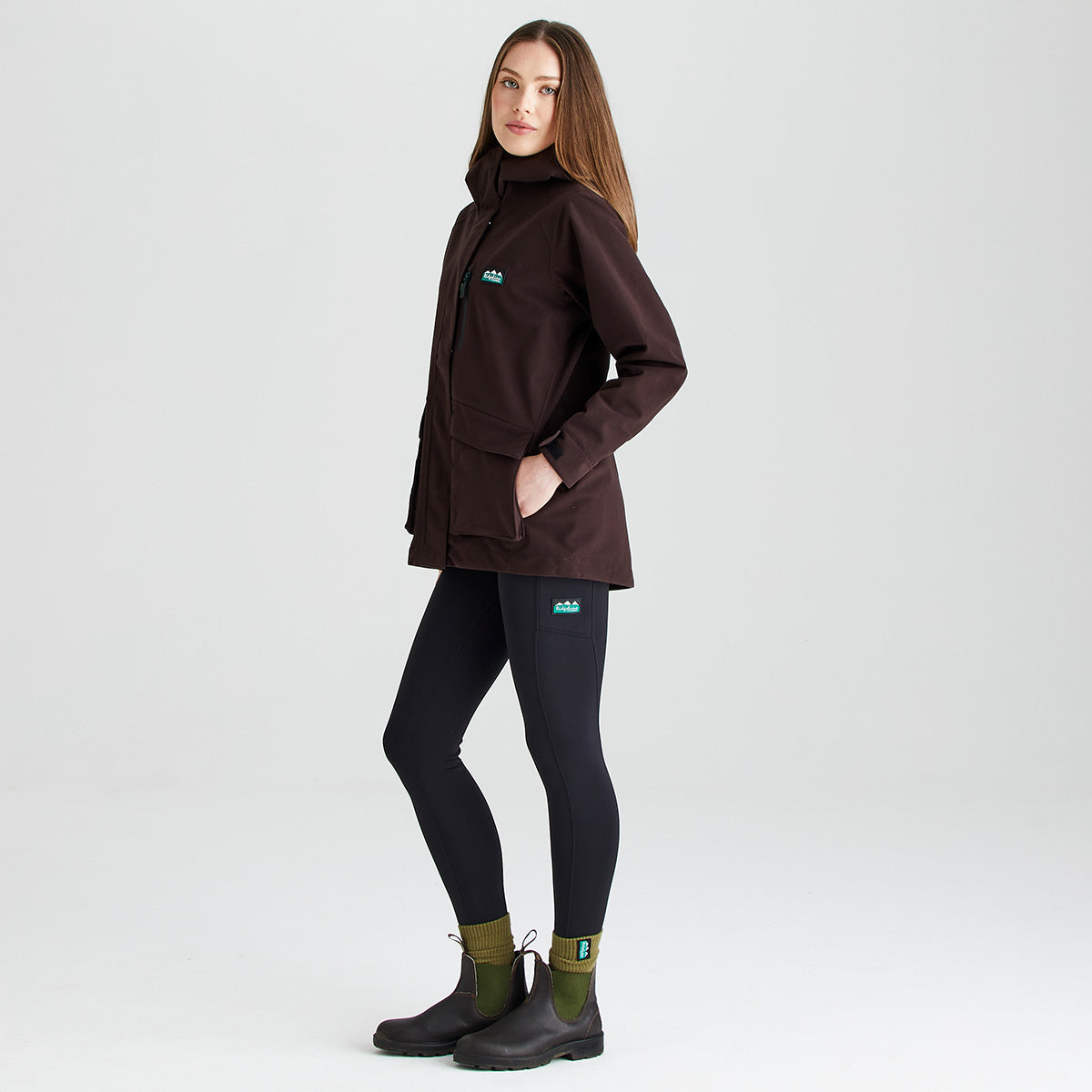 Three-quarter side view of a dark brown outdoor jacket worn with dark leggings, olive-green socks, and black ankle boots. The jacket features a high stand-up collar, a concealed full-length front zip, a vertical zipped chest pocket, and two large front flap pockets. A Ridgeline logo patch is displayed on the chest, while one hand is placed inside a pocket to demonstrate functionality. Photographed against a plain light-grey studio background, the image highlights the jacket’s relaxed fit, pr