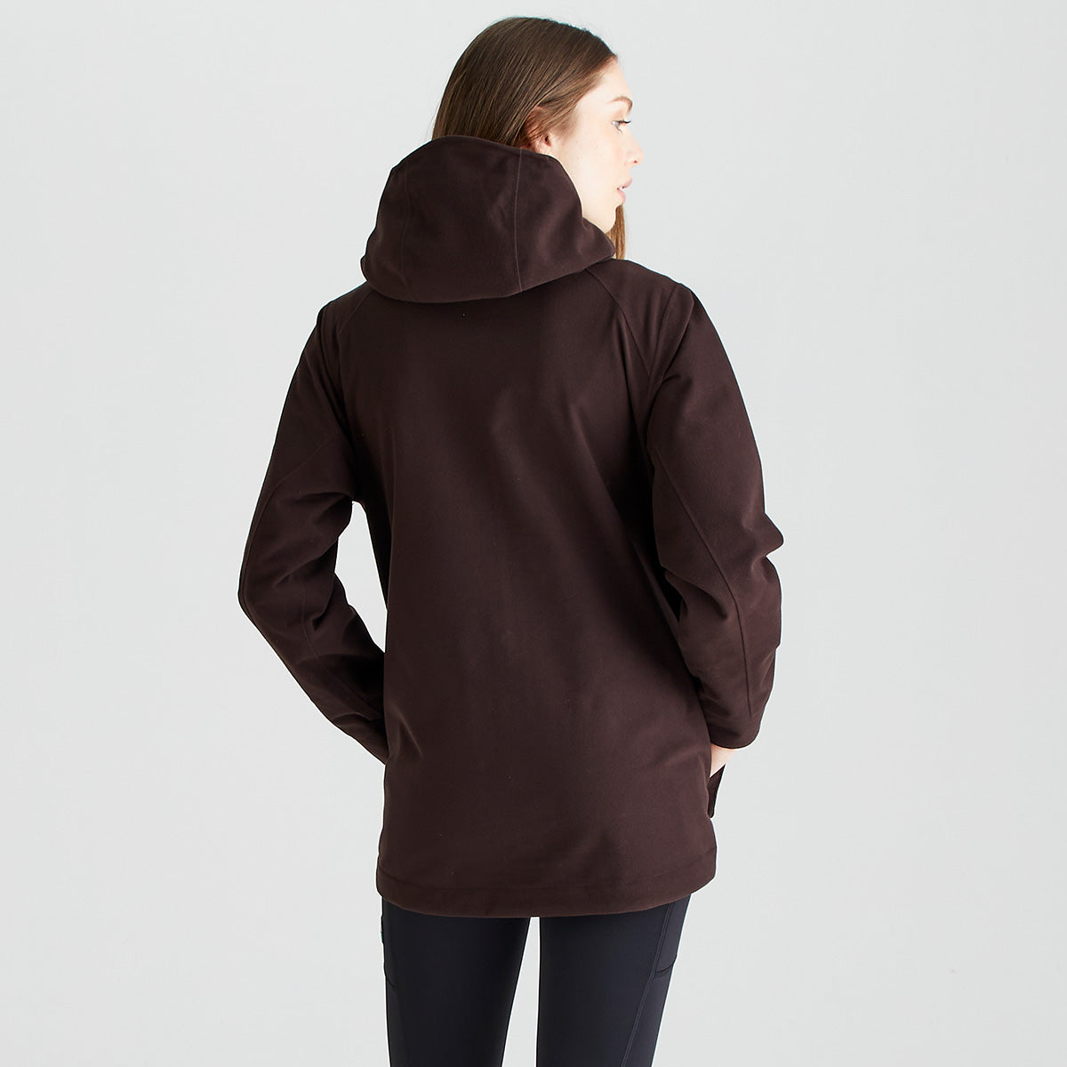 Rear view of a dark brown outdoor jacket worn with dark leggings. The jacket features a large attached hood, long sleeves, and a clean, streamlined design with minimal visible seams. The longer-length cut provides additional coverage, while the smooth outer fabric suggests a weather-resistant construction suitable for outdoor use. Photographed against a plain light-grey studio background, the image highlights the jacket’s back fit, hood design, and practical outdoor styling.
