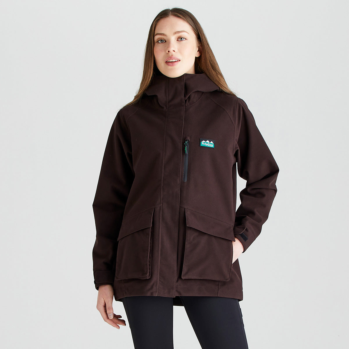 Front view of a dark brown outdoor jacket worn with dark leggings. The jacket features a high stand-up collar, a full-length front zip concealed by a storm flap, and a vertical zipped chest pocket. Two large flap-covered patch pockets are positioned at the front for storage, and a Ridgeline logo patch is displayed on the chest. The jacket has a relaxed fit with long sleeves and is photographed against a plain light-grey studio background, highlighting its practical outdoor design and weather-resistant styli
