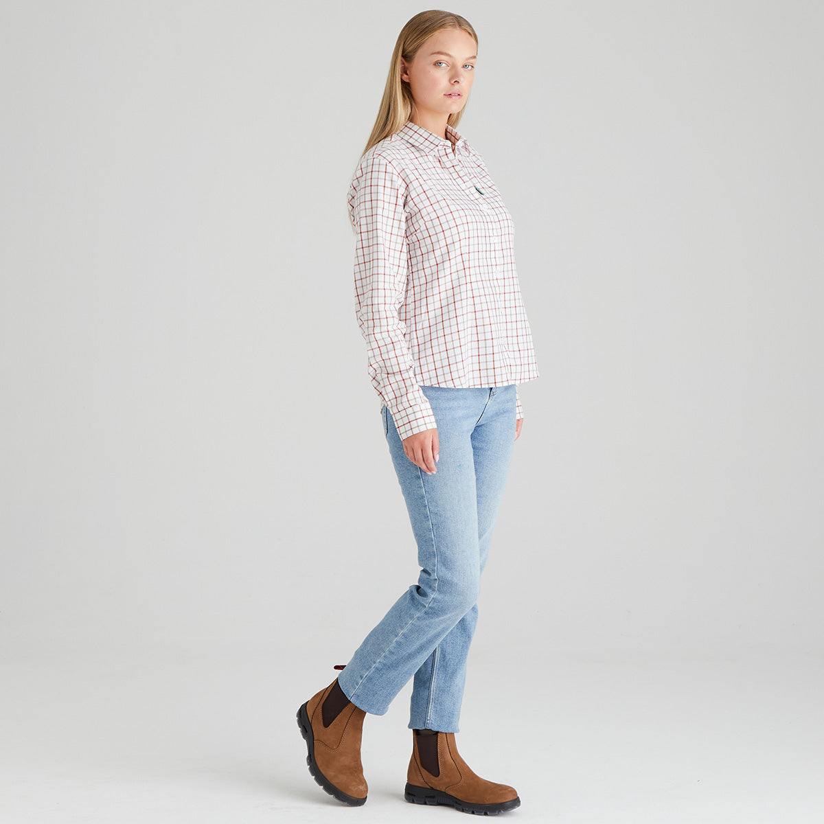 Woman wearing a plaid shirt with orange lines, jeans, and brown boots on a light grey background 