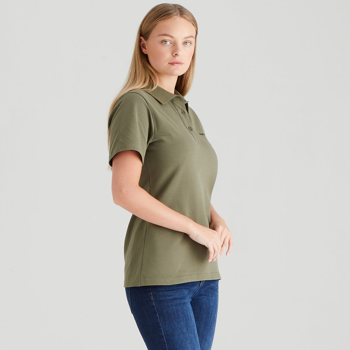 Woman wearing an olive green polo shirt and blue jeans on a light gray background