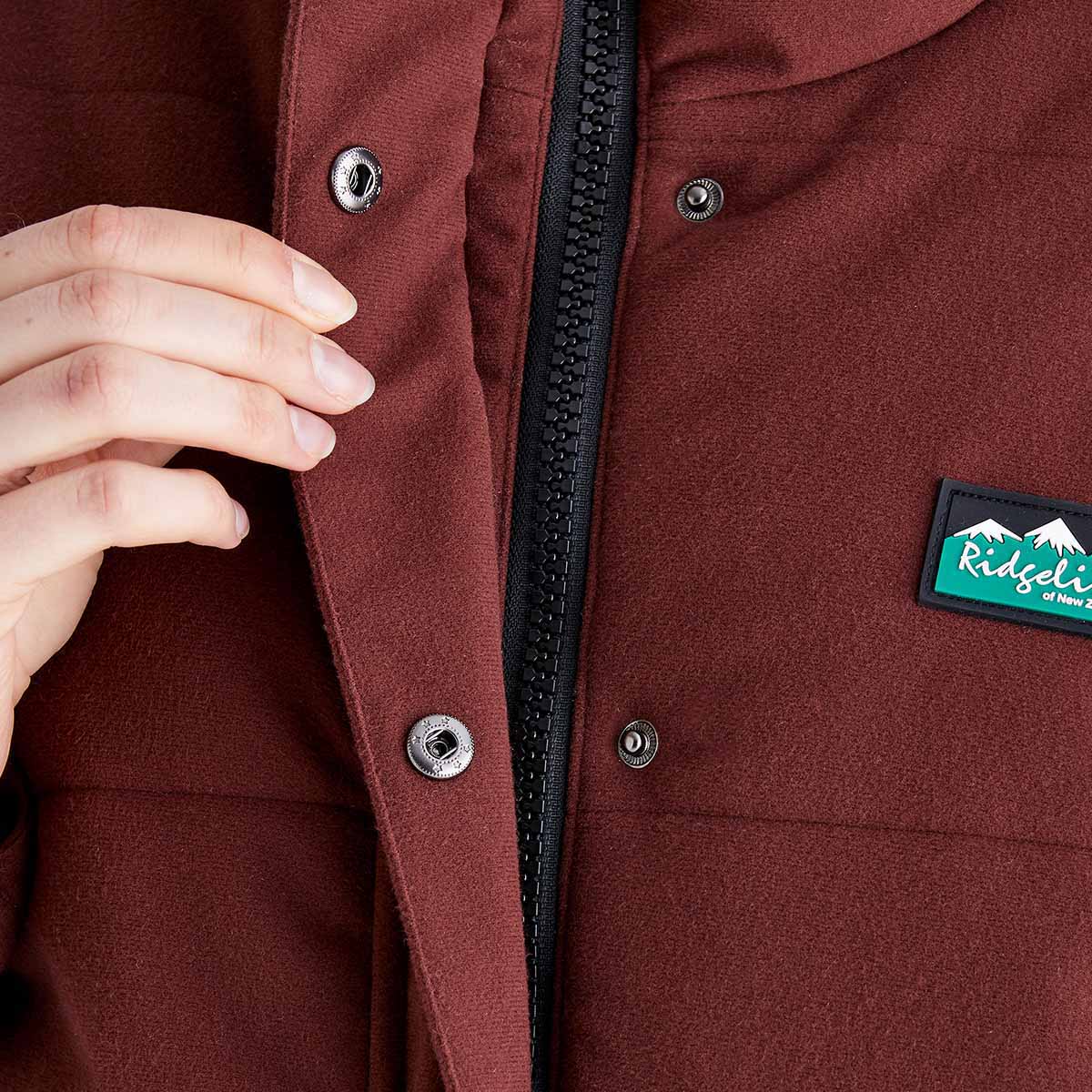 Close-up detail of a Ridgeline burgundy insulated jacket showing the full-length front zip, snap-storm flap fastening and Ridgeline chest logo patch.
