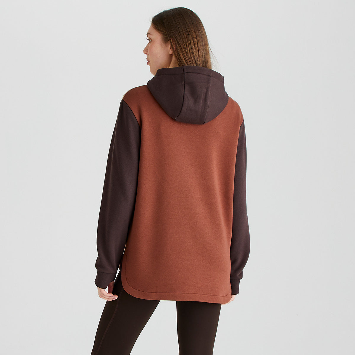 Rear view of a Ridgeline colour-block hoodie in rust brown and dark brown tones, worn with dark brown leggings. The hoodie features a large attached hood, long sleeves with ribbed cuffs, and a relaxed, longer-length fit for added coverage. The contrast dark brown hood and sleeves stand out against the rust-brown body, while the soft fleece fabric provides warmth and comfort. Photographed against a plain light-grey studio background. 