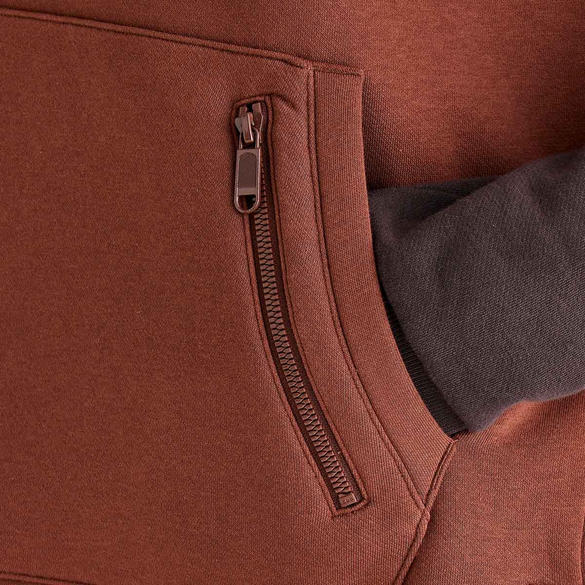 Close-up detail of a Ridgeline colour-block hoodie showing the large front kangaroo pocket and integrated vertical zipped security pocket. A hand is placed inside the pocket to demonstrate functionality, while the rust-brown fleece fabric and dark brown sleeve highlight the contrasting colour-block design. The image focuses on the hoodie’s practical storage features, durable zip fastening, and soft fleece construction.

