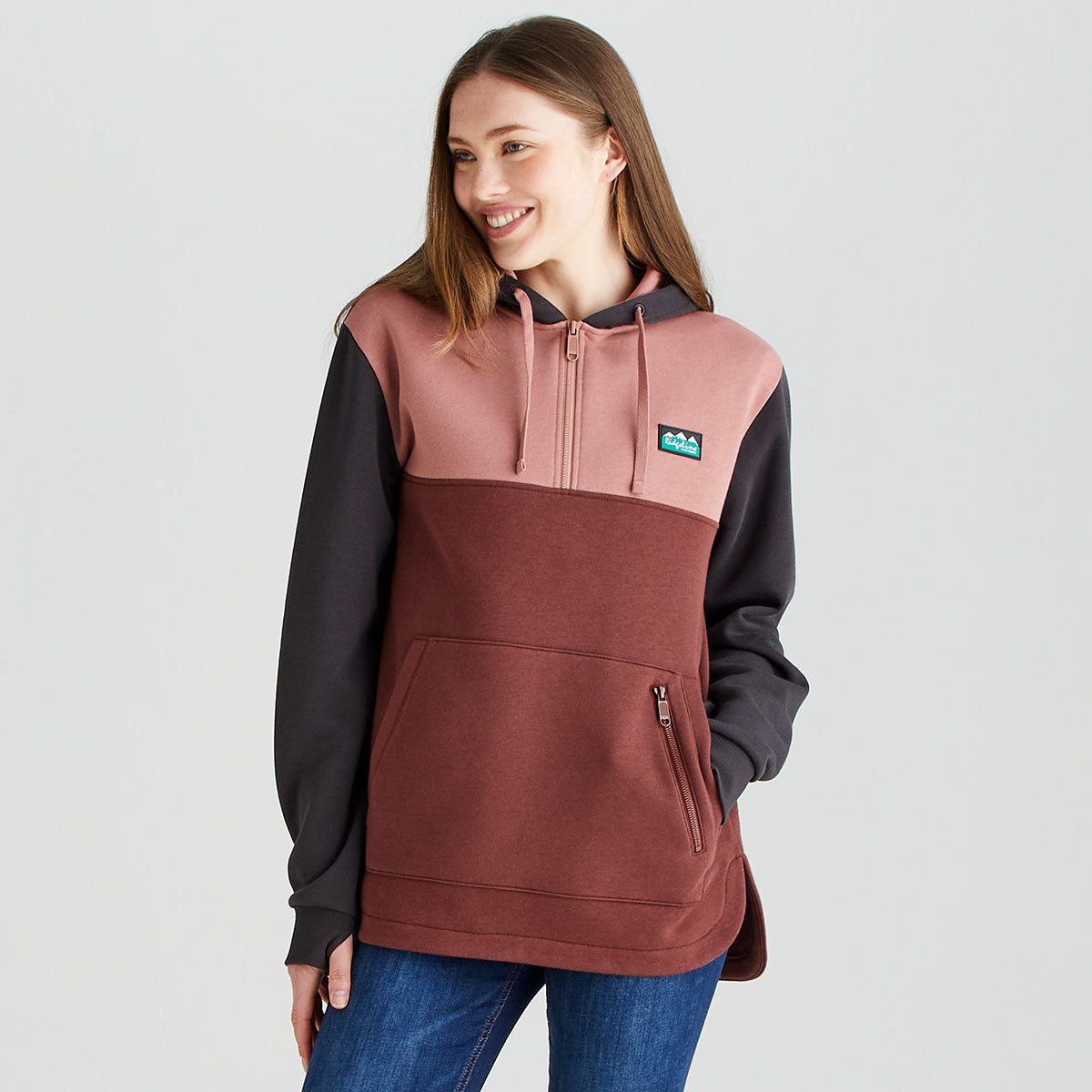 A person wearing a colour-block heavyweight Ridgeline hoodie in pink, brown and black with a 1/4 zip front, adjustable drawstring hood, kangaroo pocket and zipped security pocket, paired with blue jeans.