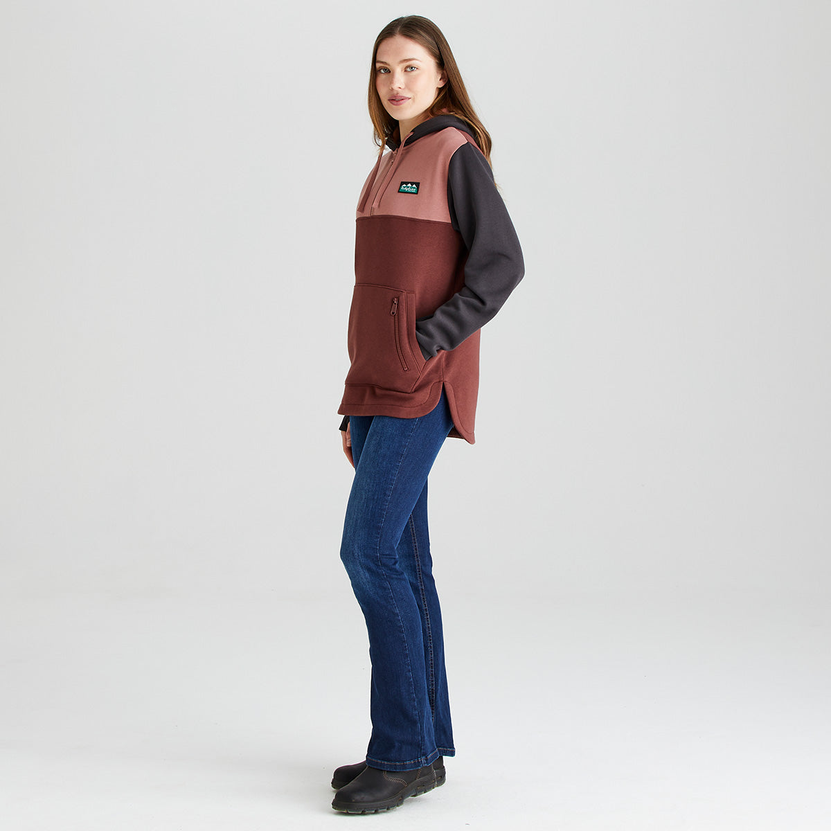 Model wearing a Ridgeline colour-block heavyweight hoodie in pink, brown and black, shown from the side with a 1/4 zip front, adjustable hood, kangaroo pocket and zipped security pocket, paired with blue jeans and outdoor boots.
