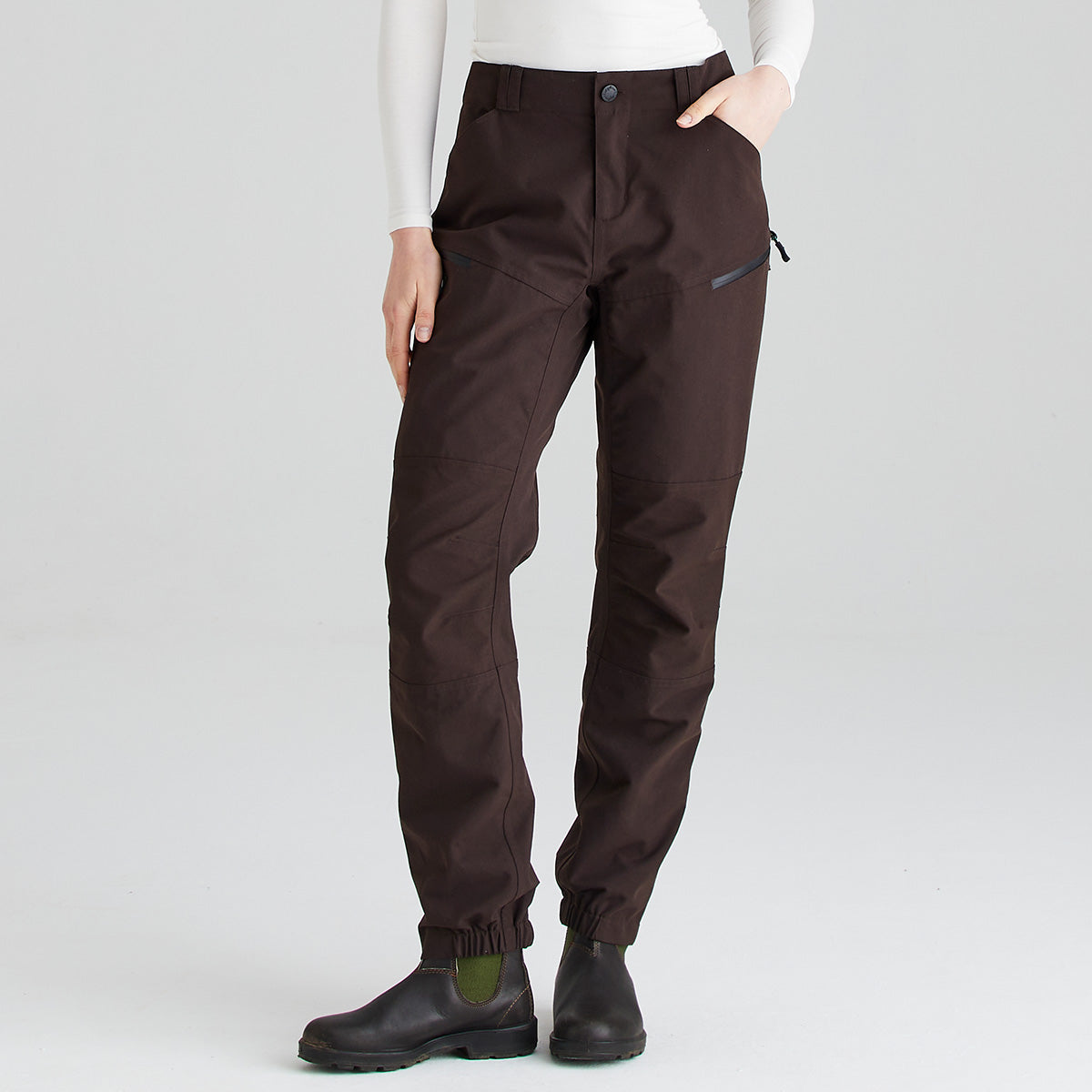Front view of dark brown waterproof trousers shown against a light gray studio background. The trousers feature a button-and-zip waistband, belt loops, articulated leg panels, and a zipped thigh pocket for secure storage. Elasticated ankle cuffs provide a close fit over footwear. The image highlights the practical design, durable construction, and streamlined fit of the trousers, paired with black rubber ankle boots.
