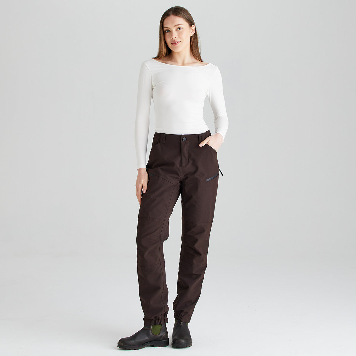 Full-length front view of dark brown waterproof trousers worn with a fitted white long-sleeve top and black ankle boots, shown against a light gray studio background. The trousers feature a button-fastening waistband, belt loops, articulated knee panels, a zipped thigh pocket, and elasticated ankle cuffs for a secure fit. The image highlights the overall fit, streamlined silhouette, and functional outdoor design of the trousers.
