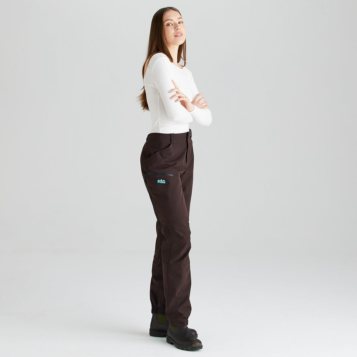 Side-profile view of dark brown waterproof trousers worn with a fitted white long-sleeve top and black ankle boots, shown against a light gray studio background. The trousers feature a button-fastening waistband, belt loops, articulated leg panels, a waterproof zipped thigh pocket, and a small logo patch on the upper leg. Elasticated ankle cuffs provide a secure fit around the boots. The image highlights the side fit, streamlined silhouette, and practical outdoor-performance features of the 