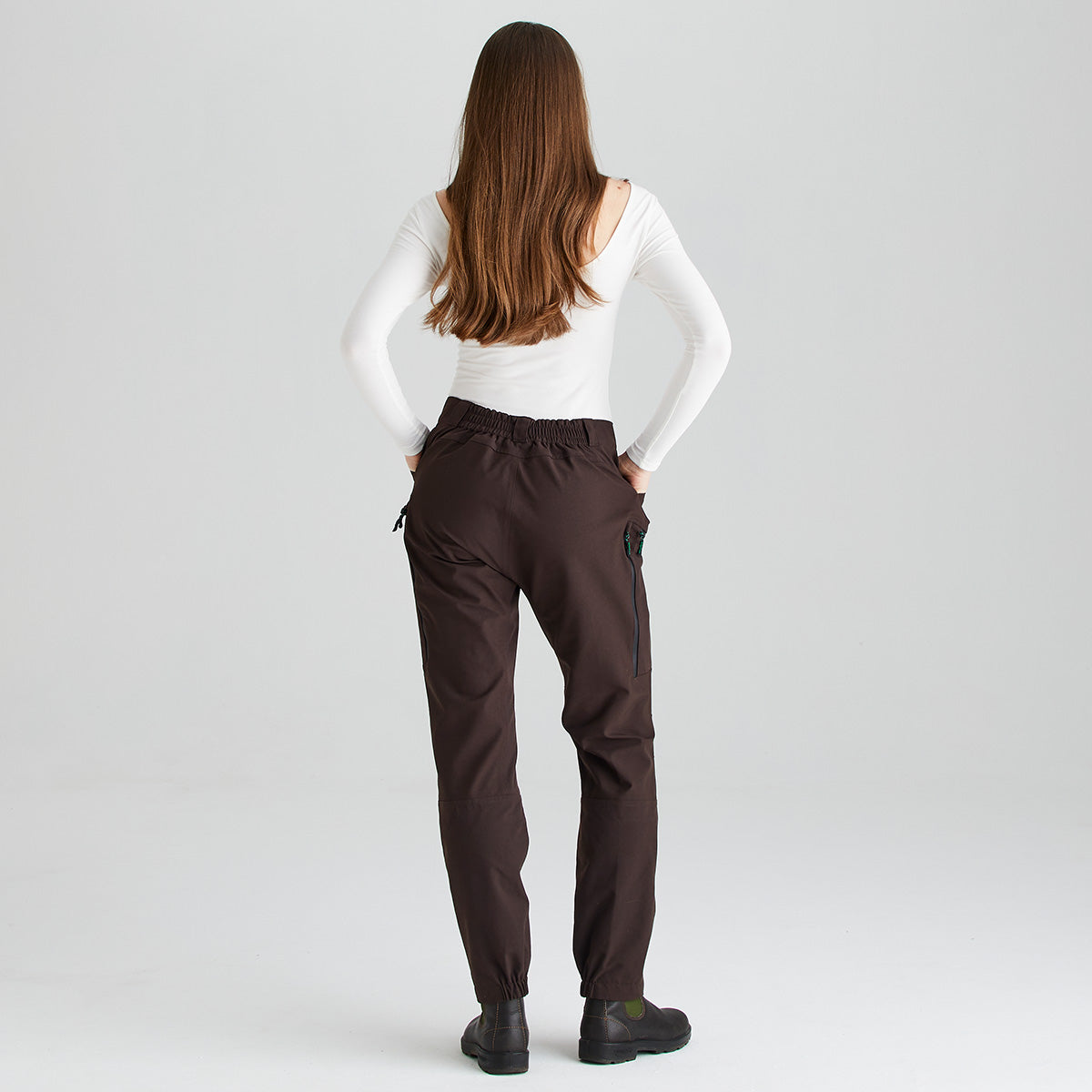 Rear full-length view of dark brown waterproof trousers worn with a fitted white long-sleeve top and black ankle boots, shown against a light gray studio background. The trousers feature an elasticated back waistband, a waterproof zipped thigh pocket, articulated leg panels, and elasticated ankle cuffs for a secure fit over footwear. The image highlights the rear fit, streamlined silhouette, and practical outdoor-performance features of the trousers.
