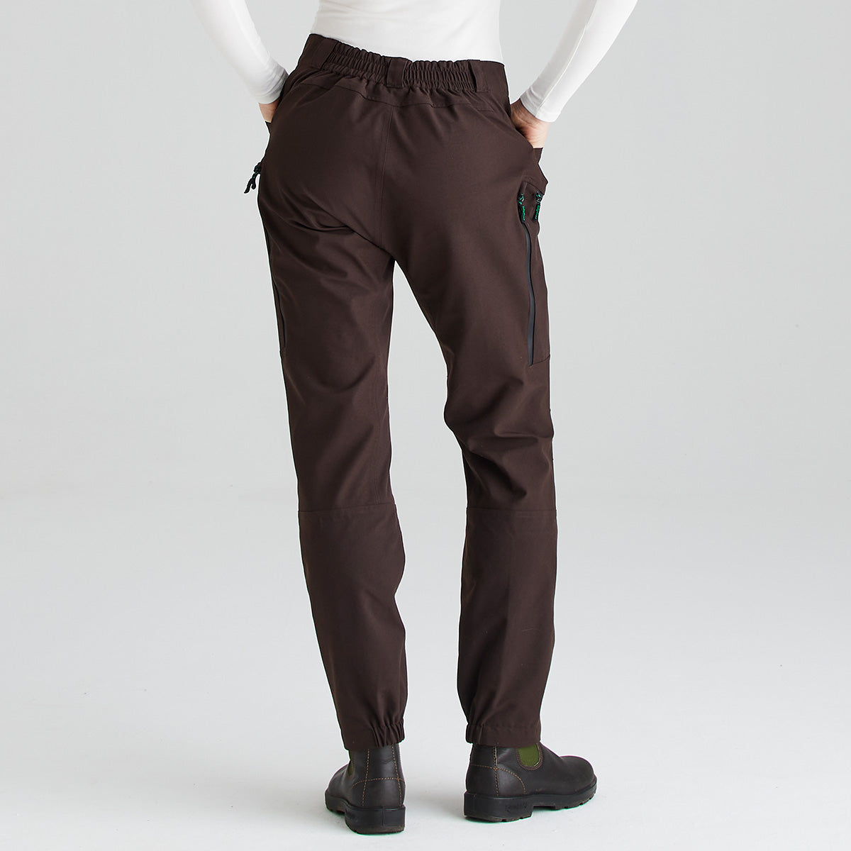 Rear view of dark brown waterproof trousers shown against a light gray studio background. The trousers feature an elasticated back waistband for added comfort, a waterproof zipped thigh pocket, articulated leg panels, and elasticated ankle cuffs that sit neatly over black ankle boots. The image highlights the back fit, durable construction, and practical outdoor-performance design of the trousers.
