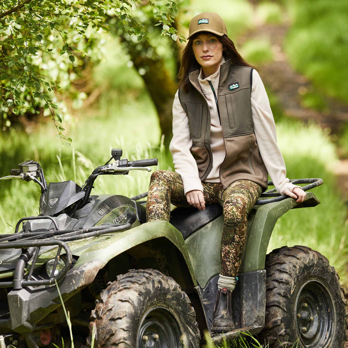 Woman wearing a A two tone brown hybrid vest with a full-length front zip,  sitting on a quad bike with trees in the background