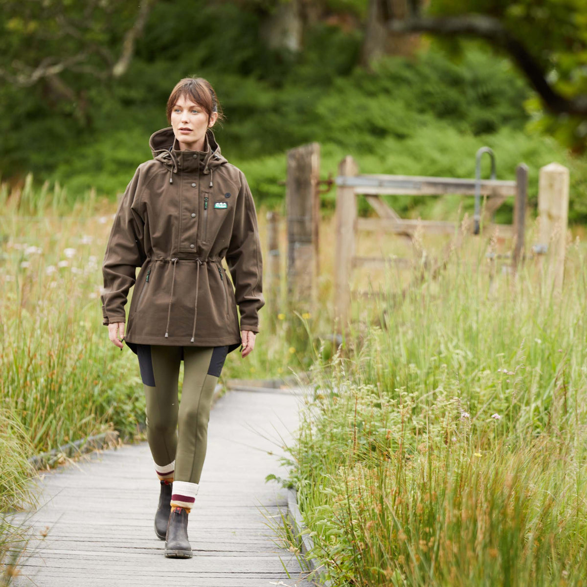 woman walking on a wooden path in a natural setting wearing a brown jacket. 