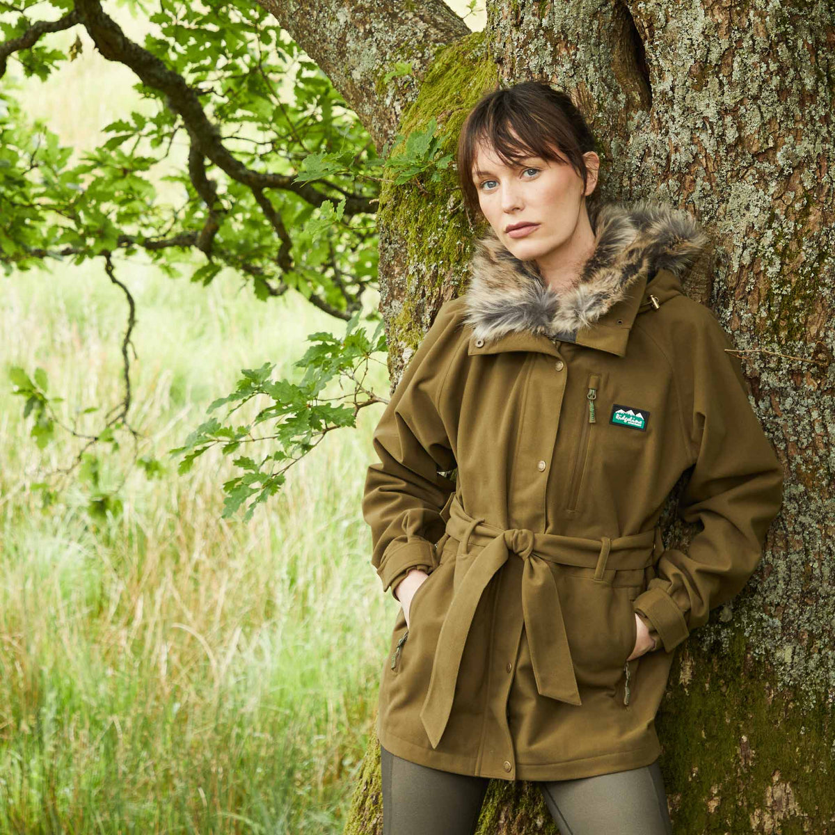 Woman wearing brown waterproof jacket leaning on a tree