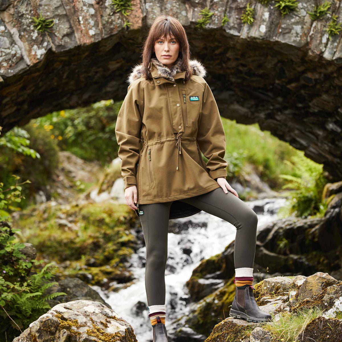 Woman wearing a beige Monsoon Nordic Smock jacket with a faux-fur collar. Standing on rocks under a bridge with water stream in the background