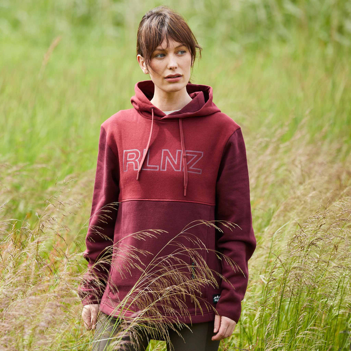 Woman wearing a two-tone red and maroon hoodie with a hood, kangaroo pocket, and the brand name 'RLNZ' printed across the chest. walking through a field of wheat