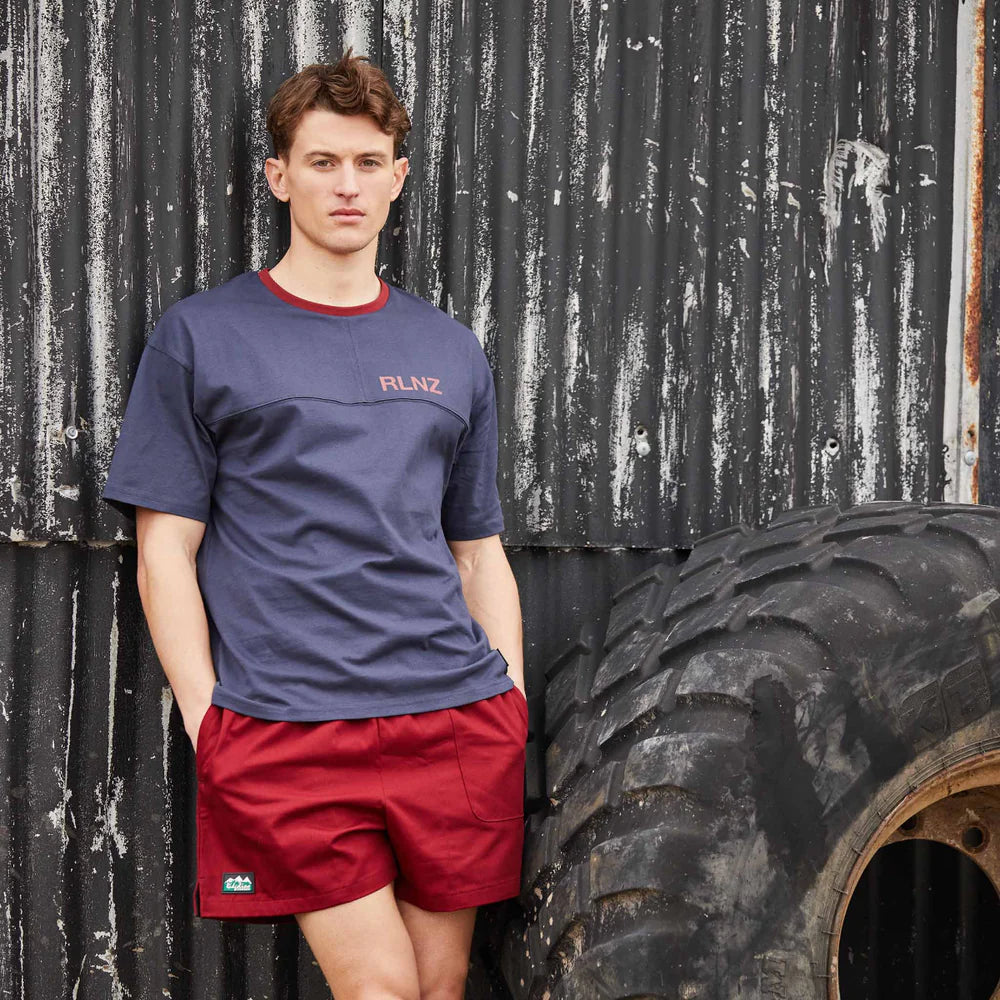 Man wearing a navy blue shirt with red trim and red shorts standing next to a large tire against a corrugated metal wall.