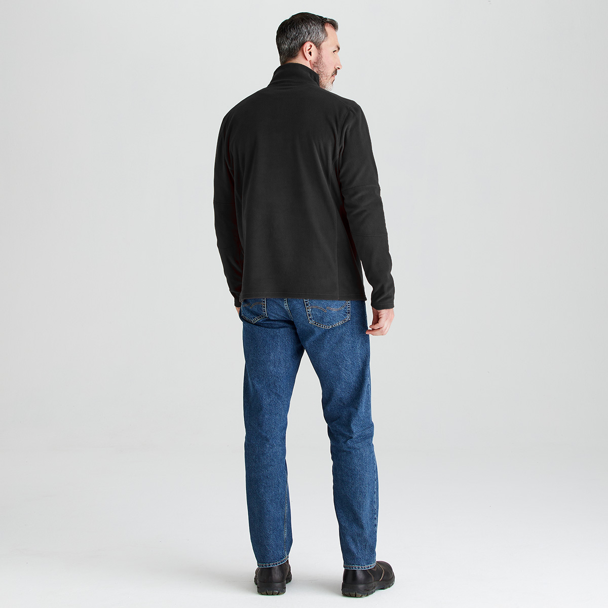 A man viewed from the back wearing a black long-sleeve quarter-zip fleece, blue jeans, and dark brown boots, standing against a light gray studio background.
