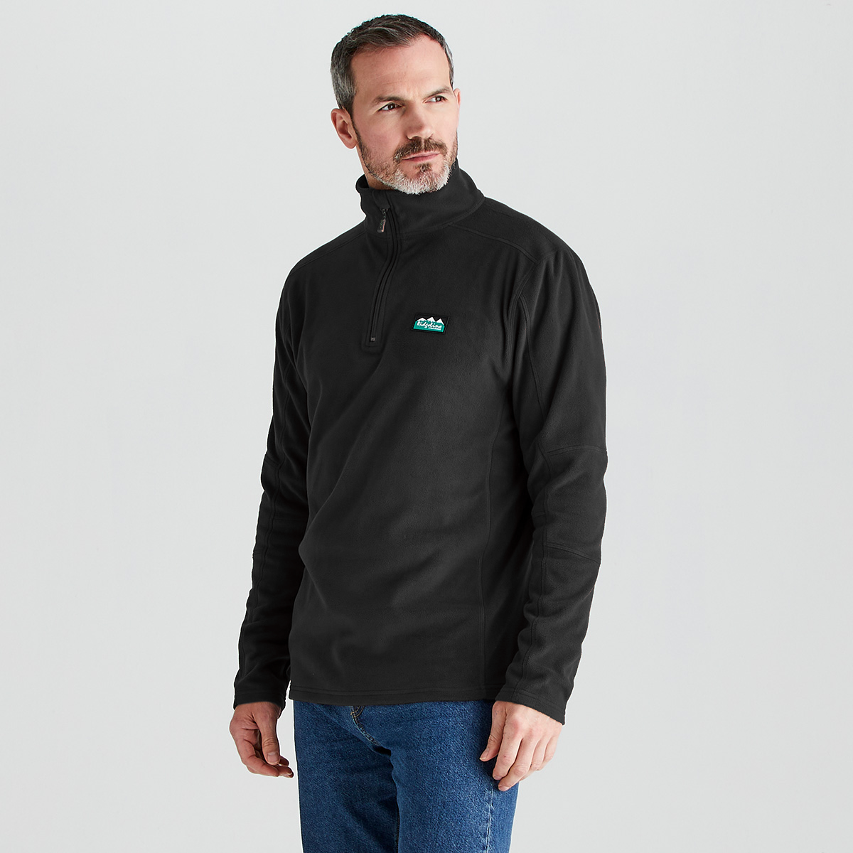 A man wearing a black quarter-zip fleece pullover with a small green and white mountain logo on the left chest, paired with blue jeans, against a light gray studio background.