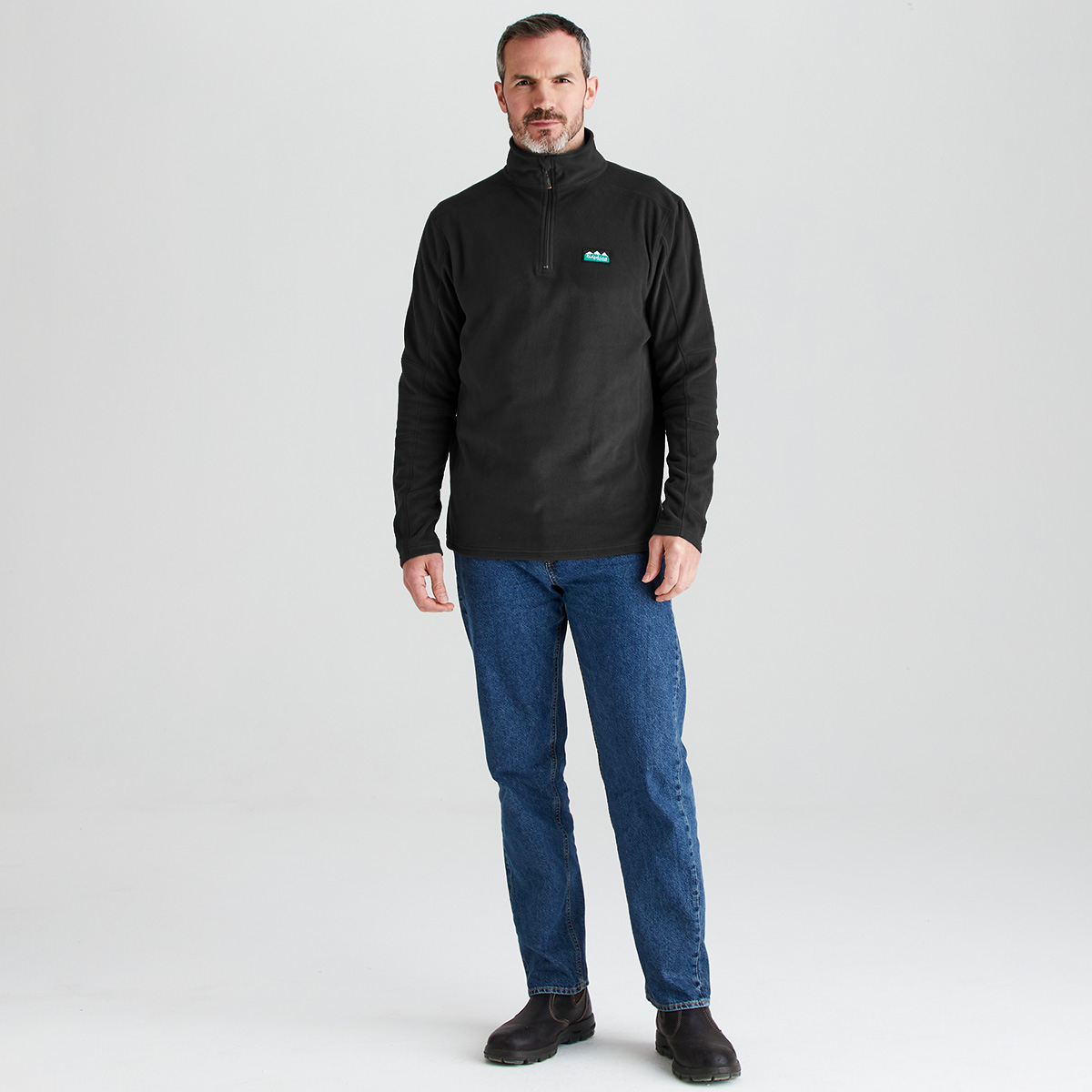 A man wearing a black quarter-zip fleece pullover with a small green and white logo on the left chest, paired with blue jeans and black boots, standing against a light gray studio background.