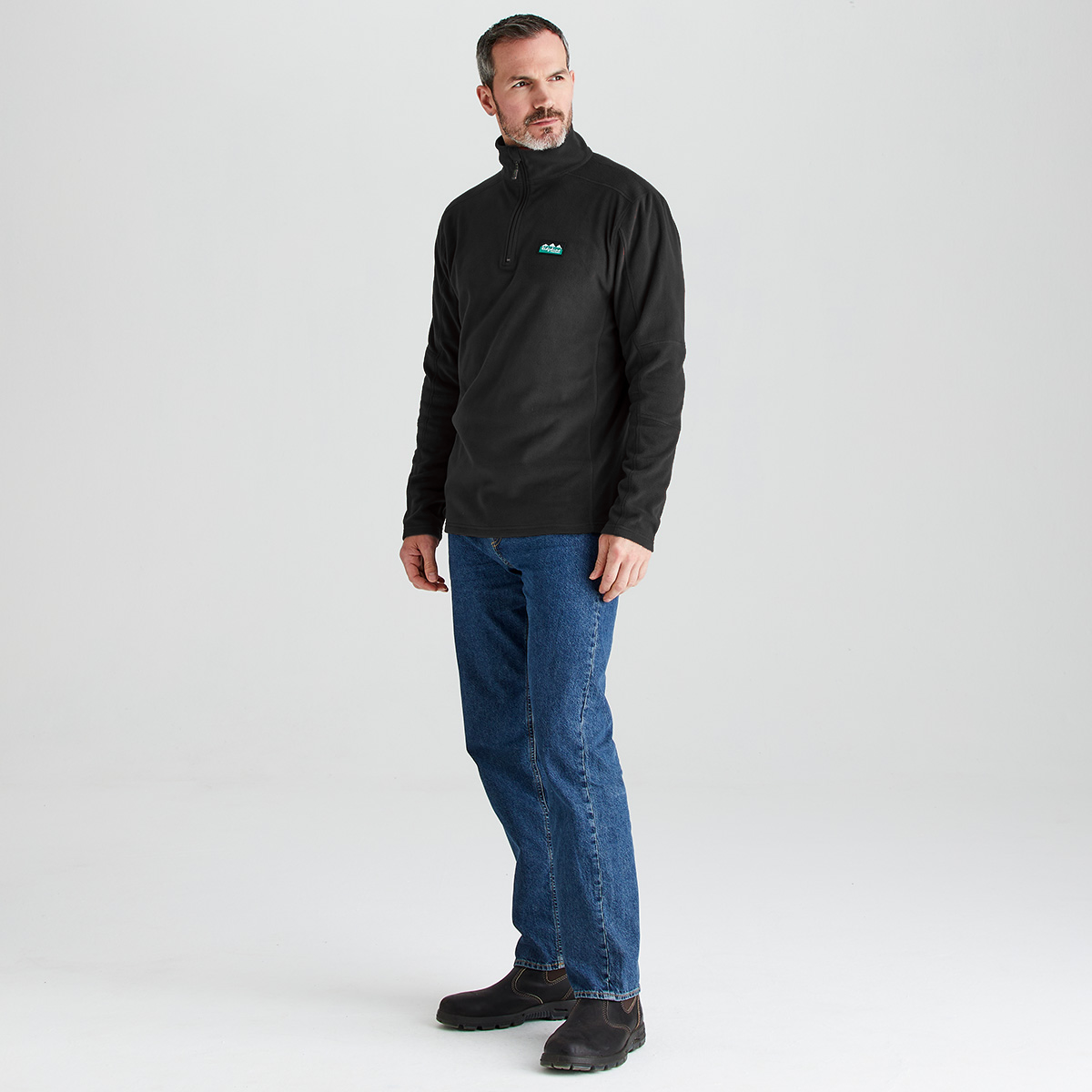 A man wearing a black quarter-zip pullover with a small teal logo on the left chest, paired with blue jeans and dark brown boots, standing against a light gray studio background.