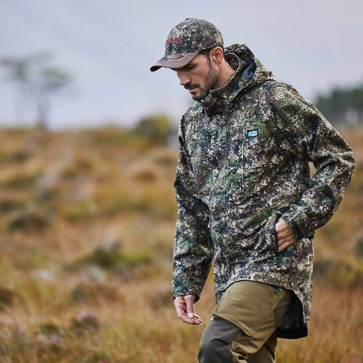 man wearing A camo green & brown coloured Ridgeline Monsoon waterproof Jacket with a detachable hood, zip front, and multiple pockets. walking through a field 